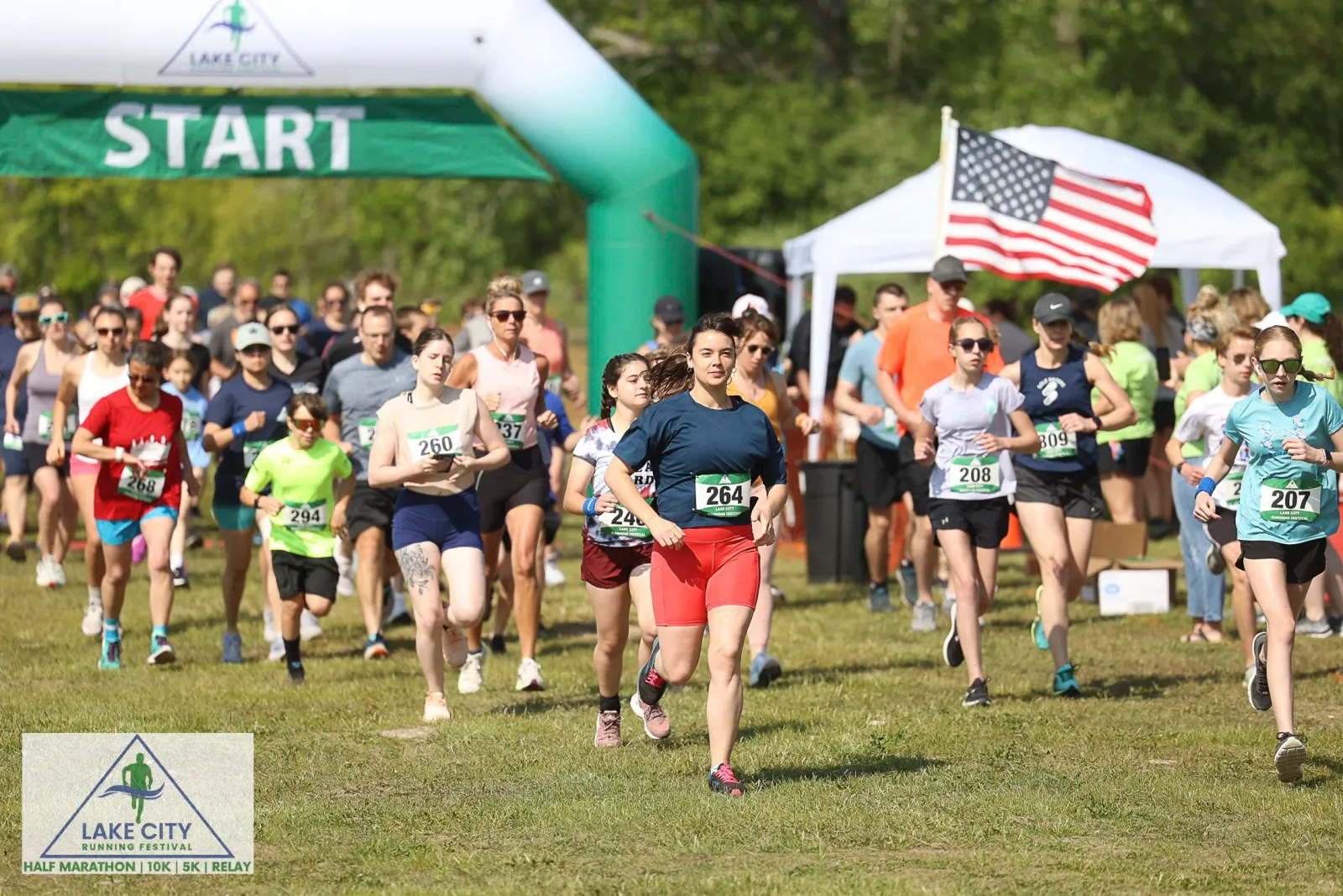Runners starting a race under a green archway with 'START' written on it, with an American flag visible in the background.