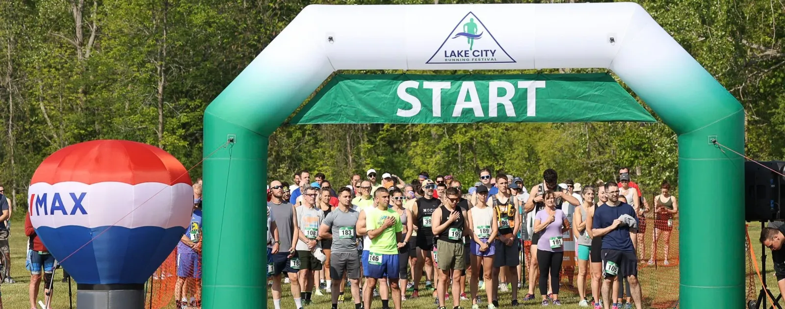 A crowd of runners stand at the start line under a green and white inflatable archway with 'START' banner.