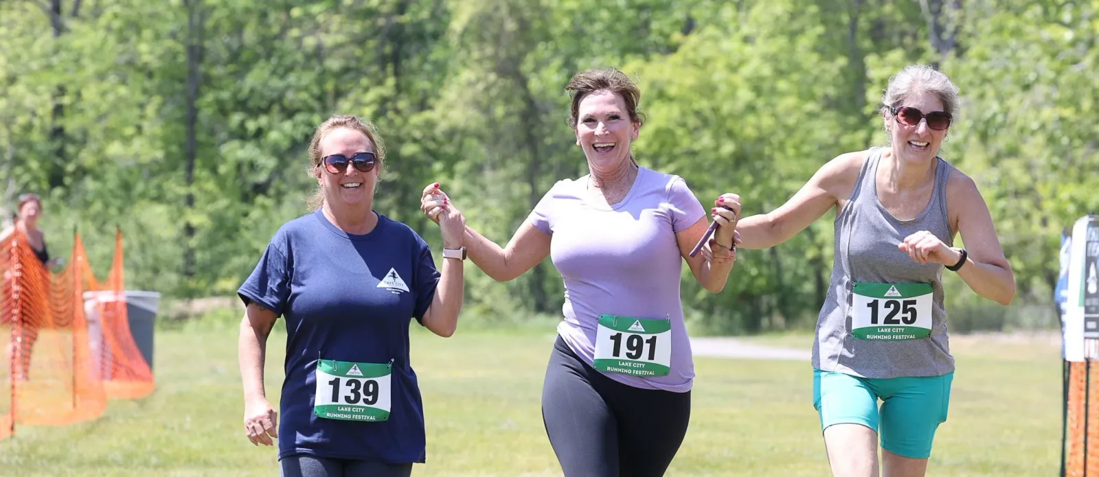 Three women running a race, smiling and holding hands, with race bibs visible.