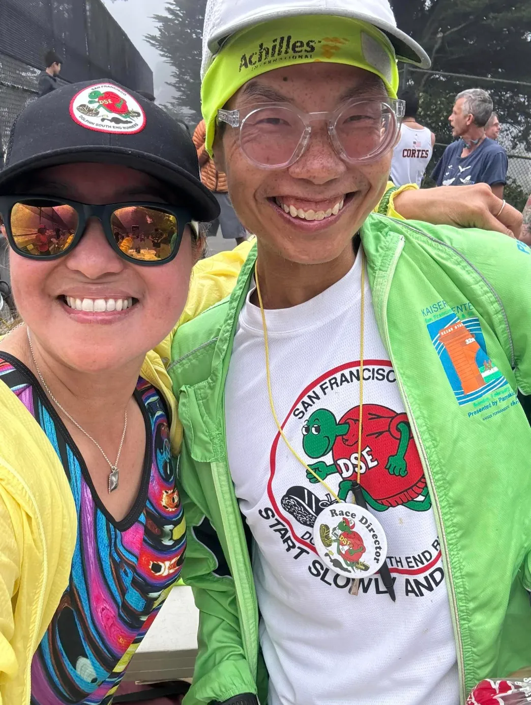 Two smiling runners, one wearing a black hat with a race logo and sunglasses, the other wearing a green jacket and a white t-shirt with a race logo, posing for a photo.