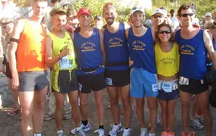 Group of runners in racing attire smiling at the camera, with race bibs visible, at the start of a race.
