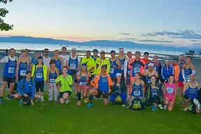 A group of runners in athletic wear pose for a photo on a grassy area with a waterfront and distant mountains in the background.