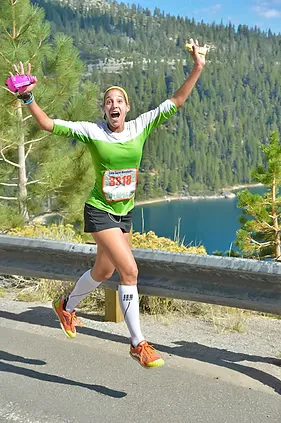 A female runner with arms raised in celebration, running on a road with a scenic view of Lake Tahoe in the background.