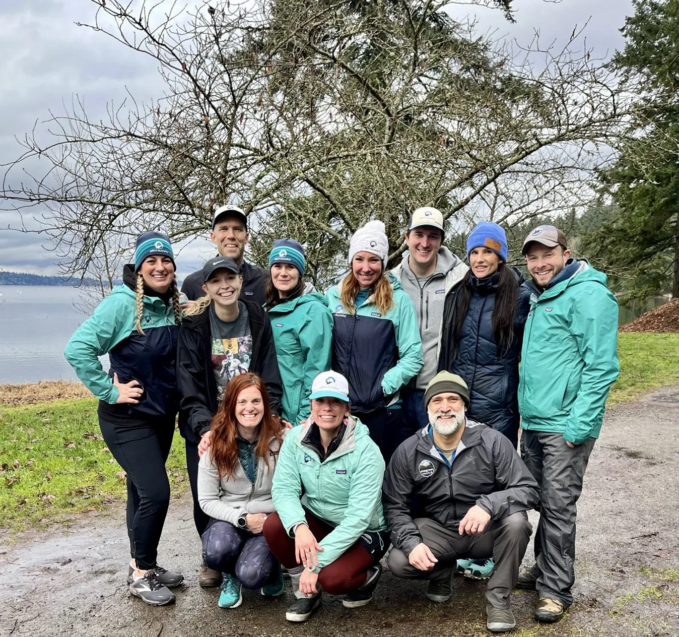 A group of ten people, some wearing matching teal jackets and hats, pose for a photo outdoors near a body of water.