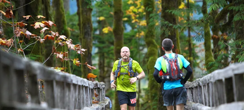 Two runners crossing a wooden bridge on a wooded trail, with autumn leaves visible.