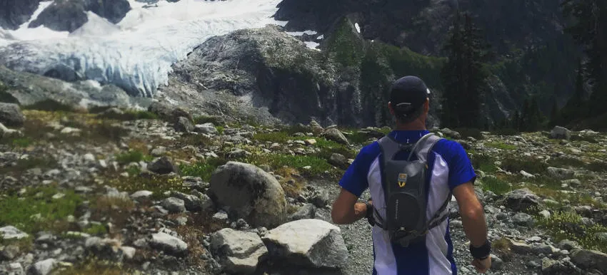 A runner with a backpack runs on a rocky mountain trail with a glacier in the background.