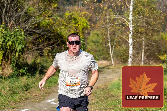 A male runner wearing sunglasses and a race bib runs on a dirt path surrounded by trees with autumn foliage.