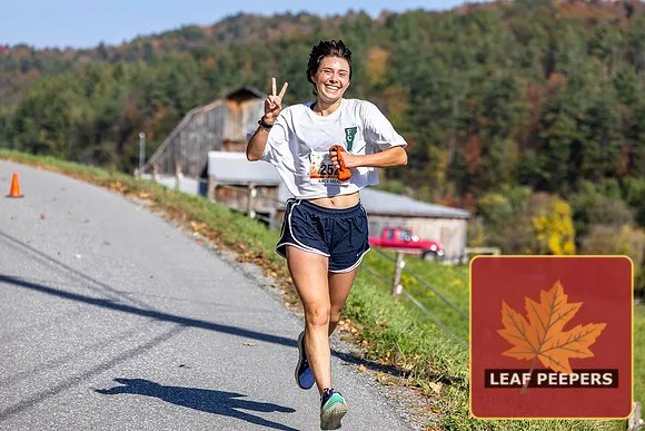 A female runner smiles and holds up two fingers in a peace sign while running on a road with a barn and autumn foliage in the background.