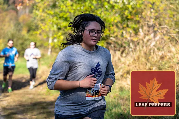 A runner with glasses and dark hair runs on a wooded path, with other runners behind her.