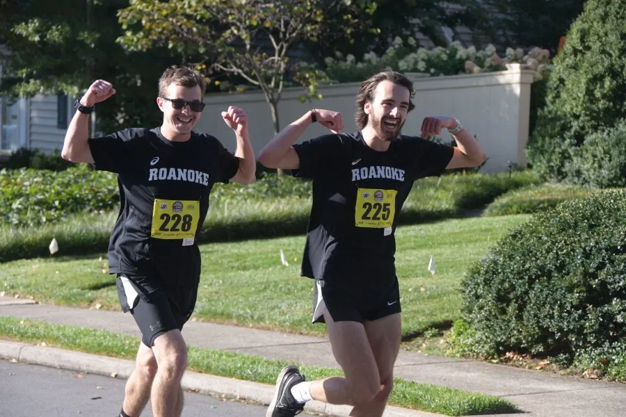 Two male runners in black shirts with 'Roanoke' and bib numbers 228 and 225, flexing their biceps and smiling as they run on a road.