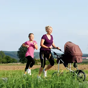 A woman and a young girl running with a baby stroller in a grassy outdoor area.