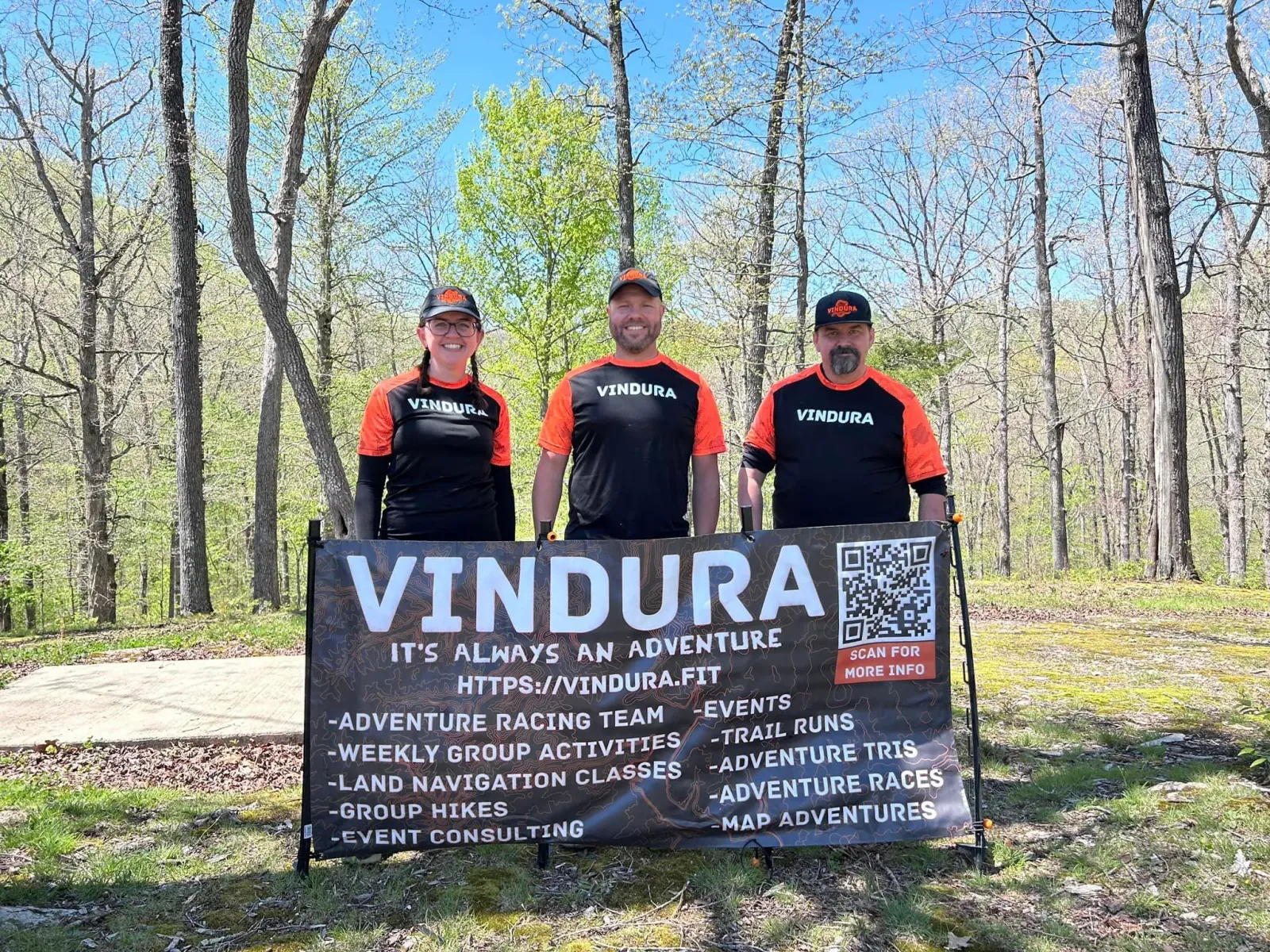 Three people wearing Vindura branded shirts and hats stand behind a banner that advertises various adventure activities including trail runs and races.