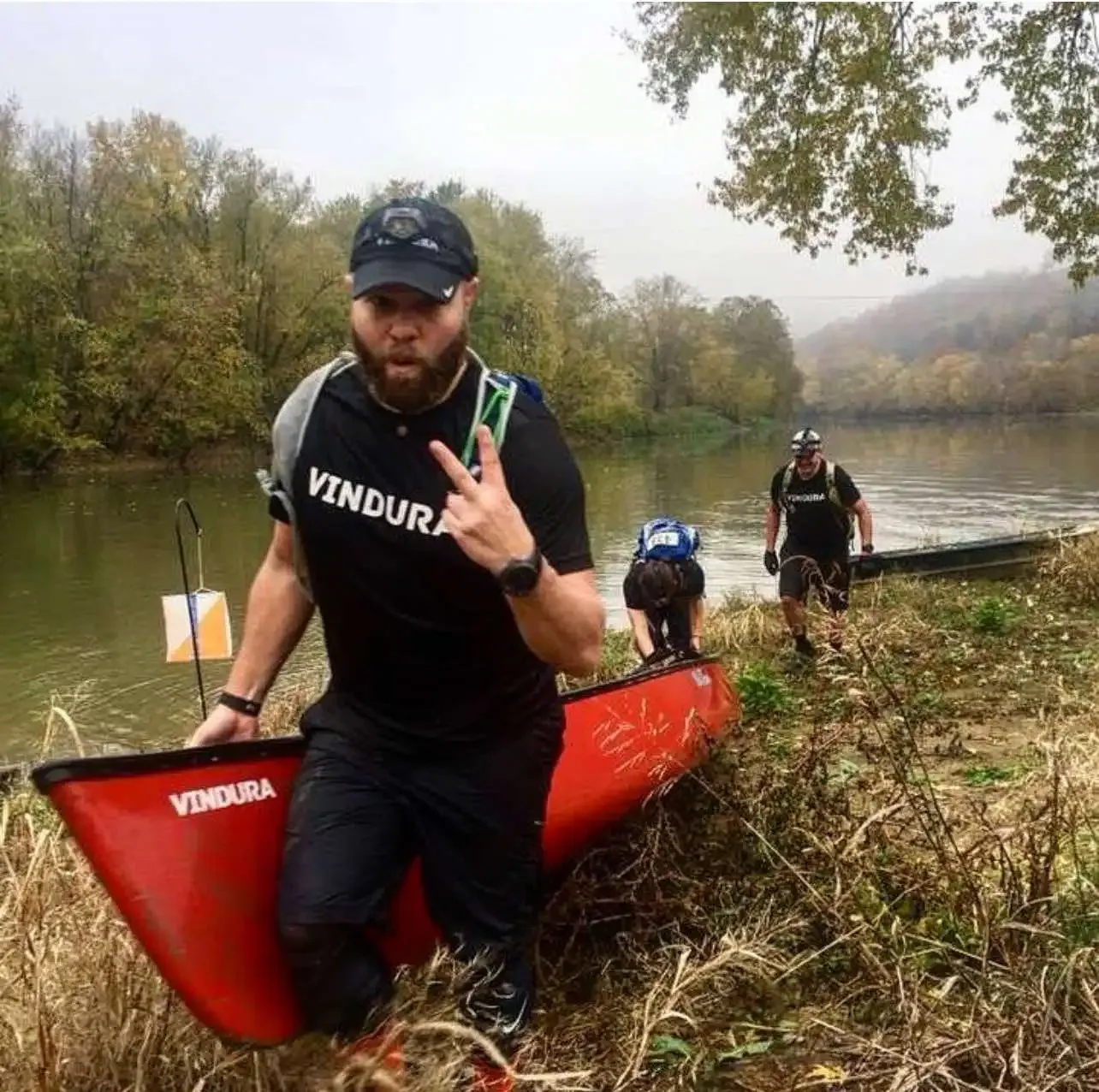 A runner in a black shirt with 'VINDURA' on it, holding a canoe and giving a peace sign, with other runners and a river in the background.