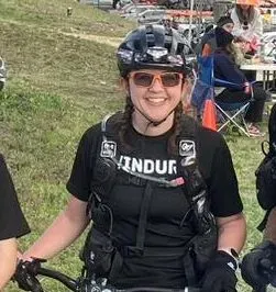 A smiling woman wearing a helmet and sunglasses, dressed in athletic gear, poses with a bicycle.