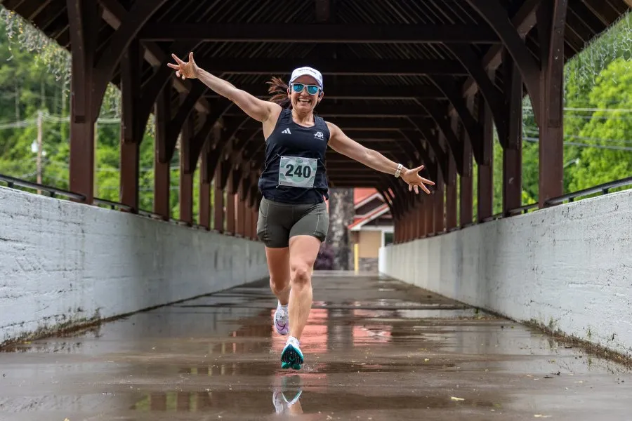 A female runner with bib number 240 runs joyfully through a covered bridge, arms outstretched.