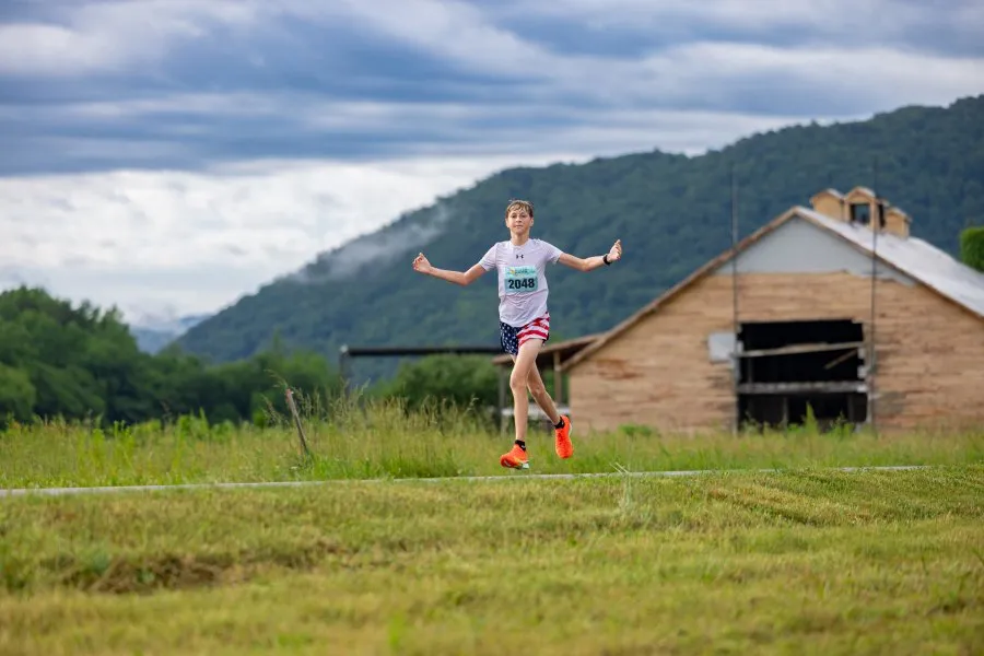 A young runner with bib number 2048 runs on a road with a barn and mountains in the background.