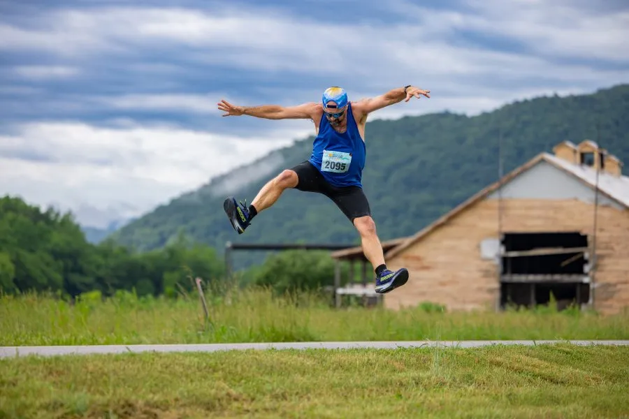 A male runner in a blue tank top and shorts jumps in the air with arms outstretched, wearing a race bib number 2095. He is in front of a rural landscape with a barn and mountains under a cloudy sky.
