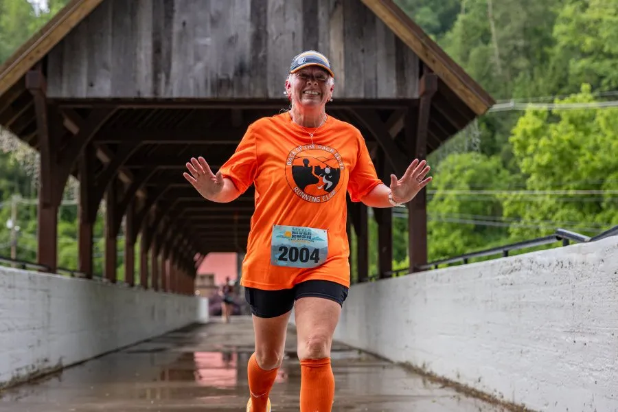 A female runner wearing an orange shirt and bib number 2004 smiles and waves as she runs across a covered bridge during the Little River Rush Half Marathon.