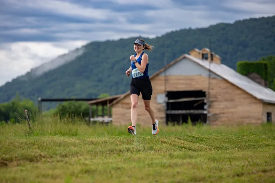 A female runner smiles as she runs past a rustic barn with a mountain in the background.