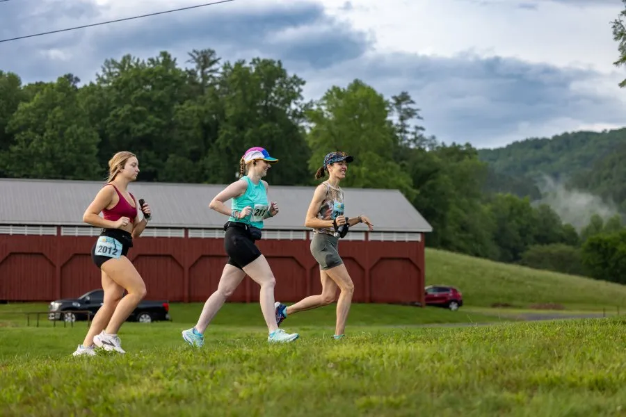 Three female runners in racing attire are running on a grassy path with a red barn and rolling hills in the background.