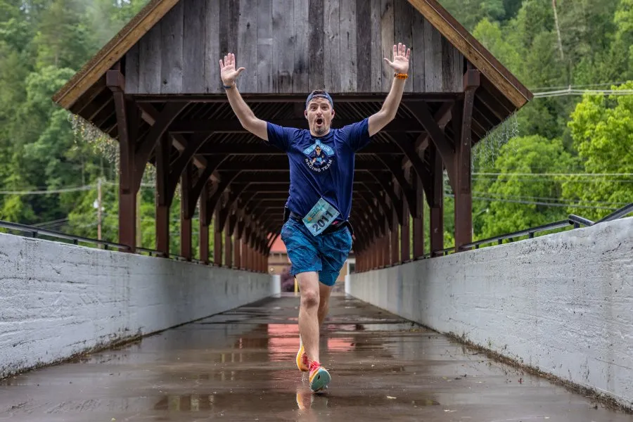 A runner with arms raised in celebration runs across a covered bridge, wearing a blue race t-shirt and blue shorts.