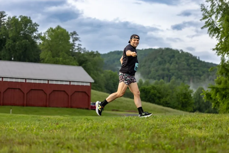 A male runner in a black t-shirt and patterned shorts runs across a grassy field with a red barn and tree-covered hills in the background.