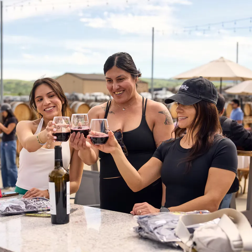 Three women toasting with glasses of red wine at an outdoor event, possibly a winery, with barrels in the background.