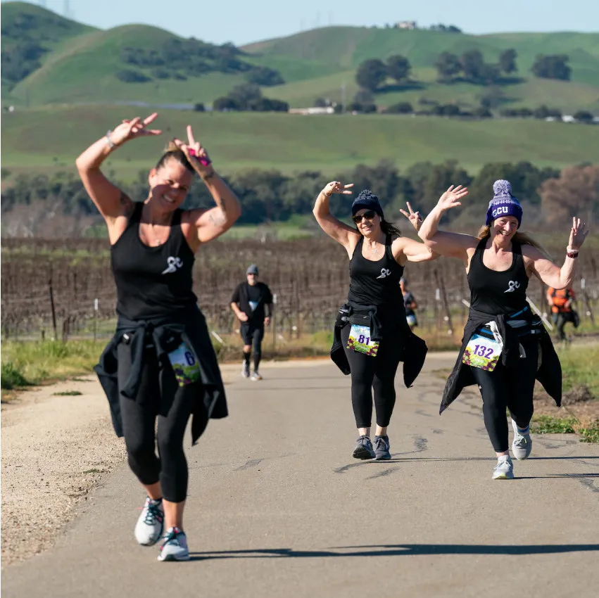 Three women running a race, two in the foreground with arms raised in a peace sign, smiling, with rolling hills and vineyards in the background.