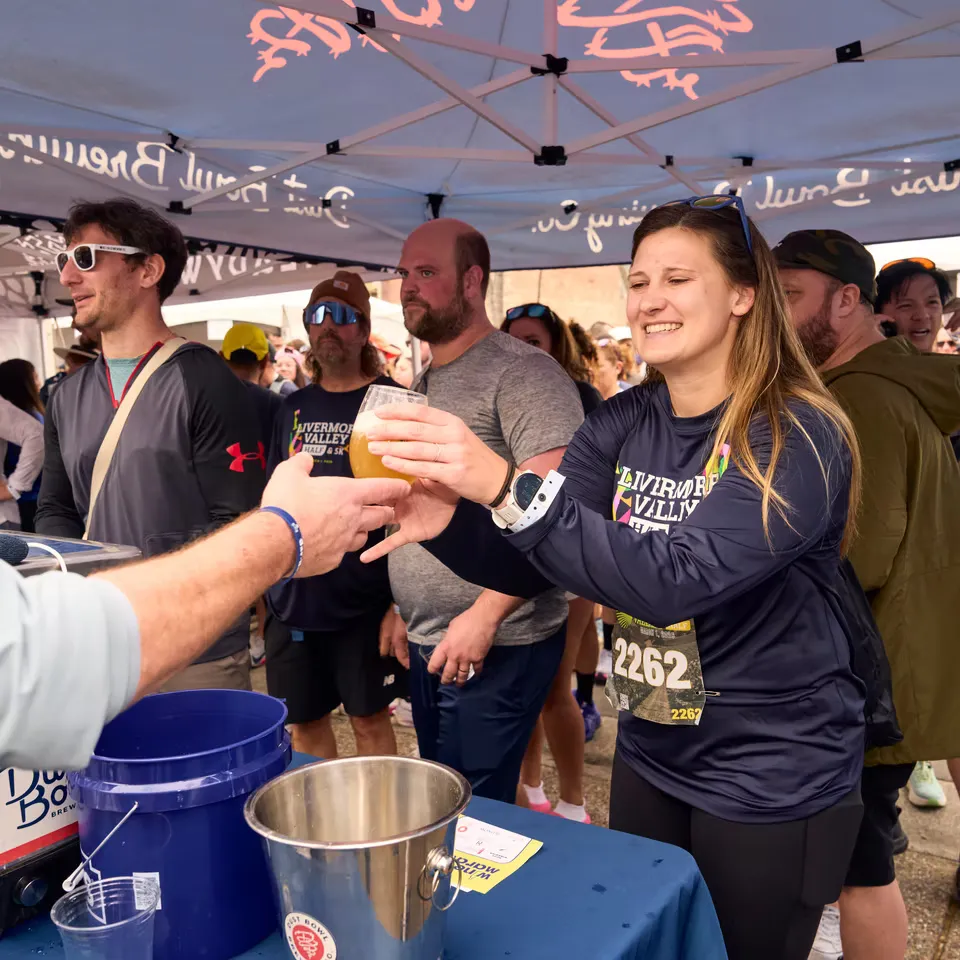 A woman in a dark blue long-sleeved shirt with a race bib showing '2262' receives a glass of beer from a server under a tent, with other runners in the background.