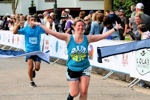 A female runner with her arms outstretched crosses the finish line with a big smile, while a male runner behind her also celebrates.