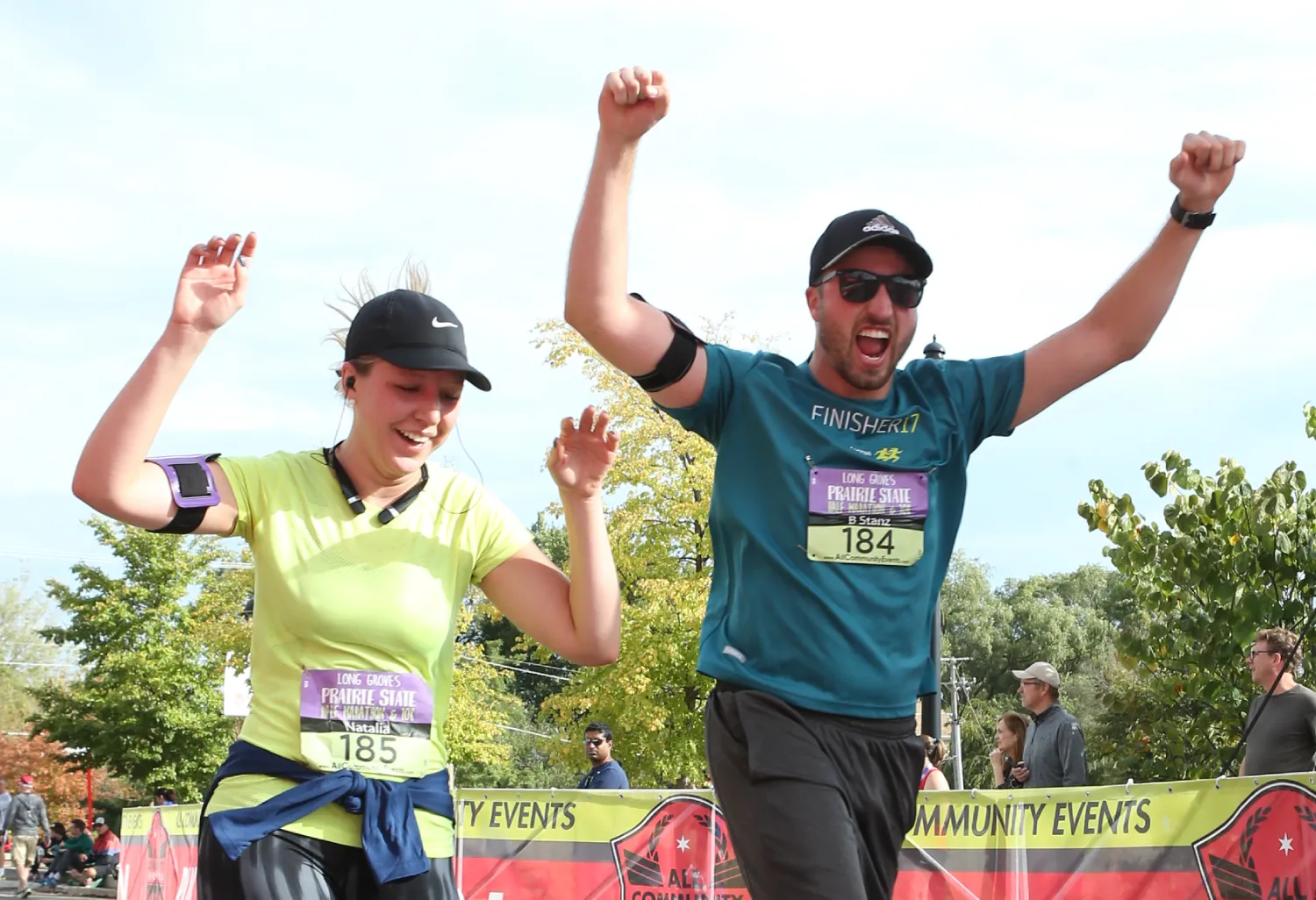 Two runners, a woman in a lime green shirt and a man in a teal shirt, celebrate with their arms raised in victory at the finish line of the Long Grove Oktoberfest Prairie State Half Marathon.