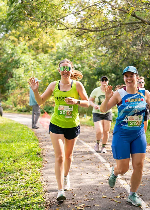 Two female runners smiling and waving while running on a paved path lined with trees.
