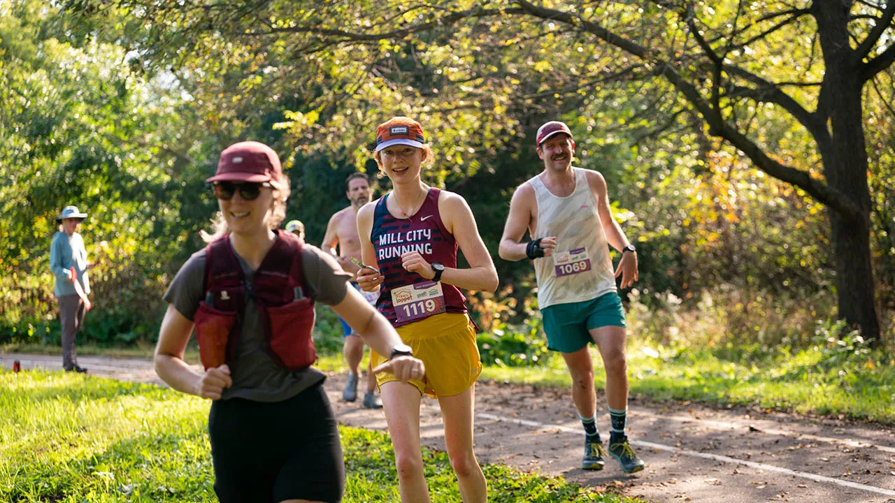 Runners on a wooded trail, with the lead runner wearing a maroon tank top that says 'MILL CITY RUNNING'.