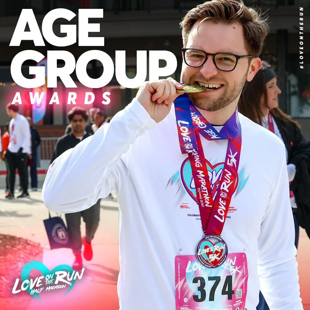 A smiling man wearing glasses and a white long-sleeved shirt, biting on a gold medal with 'Love On The Run 5K' ribbons. He has a race bib with the number 374. The text 'AGE GROUP AWARDS' is prominently displayed at the top of the image.