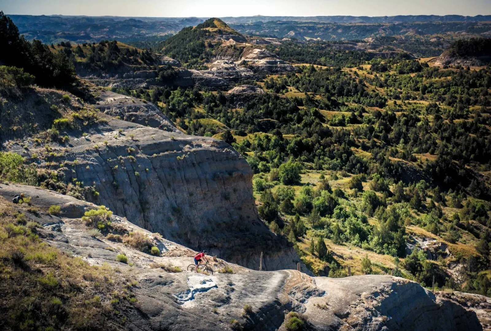 A lone cyclist navigates a rocky trail through a scenic, hilly landscape with scattered trees.