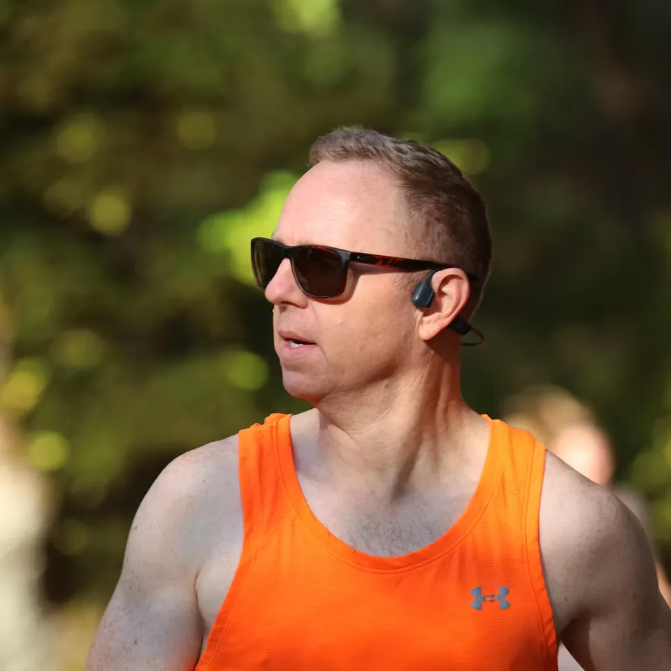 A male runner wearing sunglasses and an orange tank top, running outdoors.