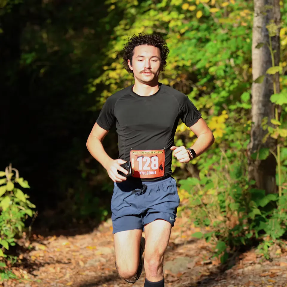 A male runner with curly hair and a mustache runs on a wooded trail, wearing bib number 128.