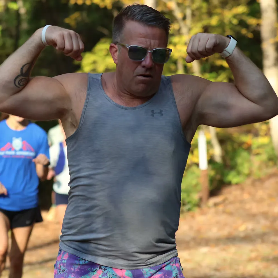 A man in a grey tank top and colorful shorts flexes his biceps while running on a trail, with other runners blurred in the background.