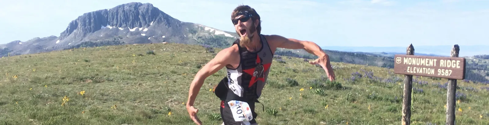 A runner with an excited expression runs on a mountain trail, with a sign indicating Monument Ridge and an elevation of 9587 feet.