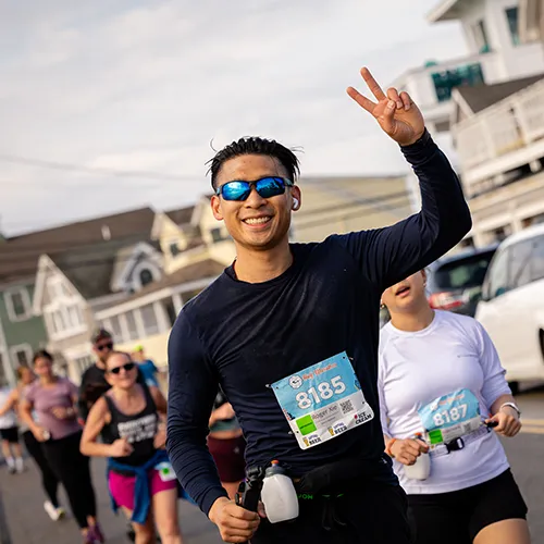 A male runner smiles and gives a peace sign while running in a race, with other runners in the background.