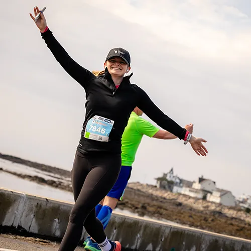 A female runner with arms outstretched in celebration, wearing a black long-sleeved shirt and a race bib, with a scenic waterfront in the background.