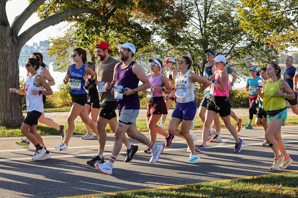 A group of runners race along a paved path with trees and water in the background.