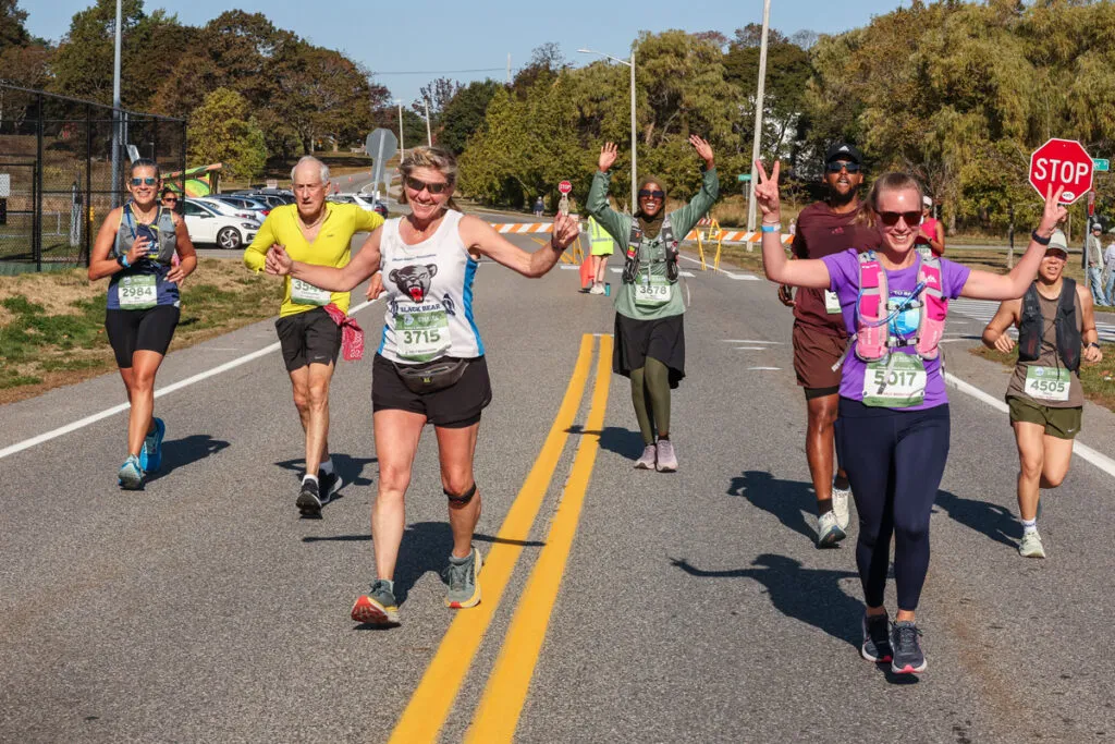 A group of runners smiles and celebrates as they run down a road, with some raising their arms in victory.