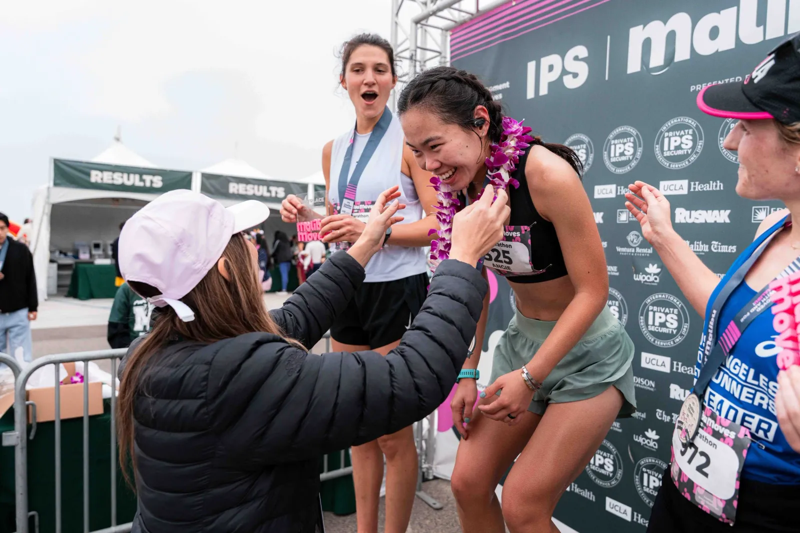 A runner receives a lei after crossing the finish line, with other runners and a celebratory atmosphere in the background.