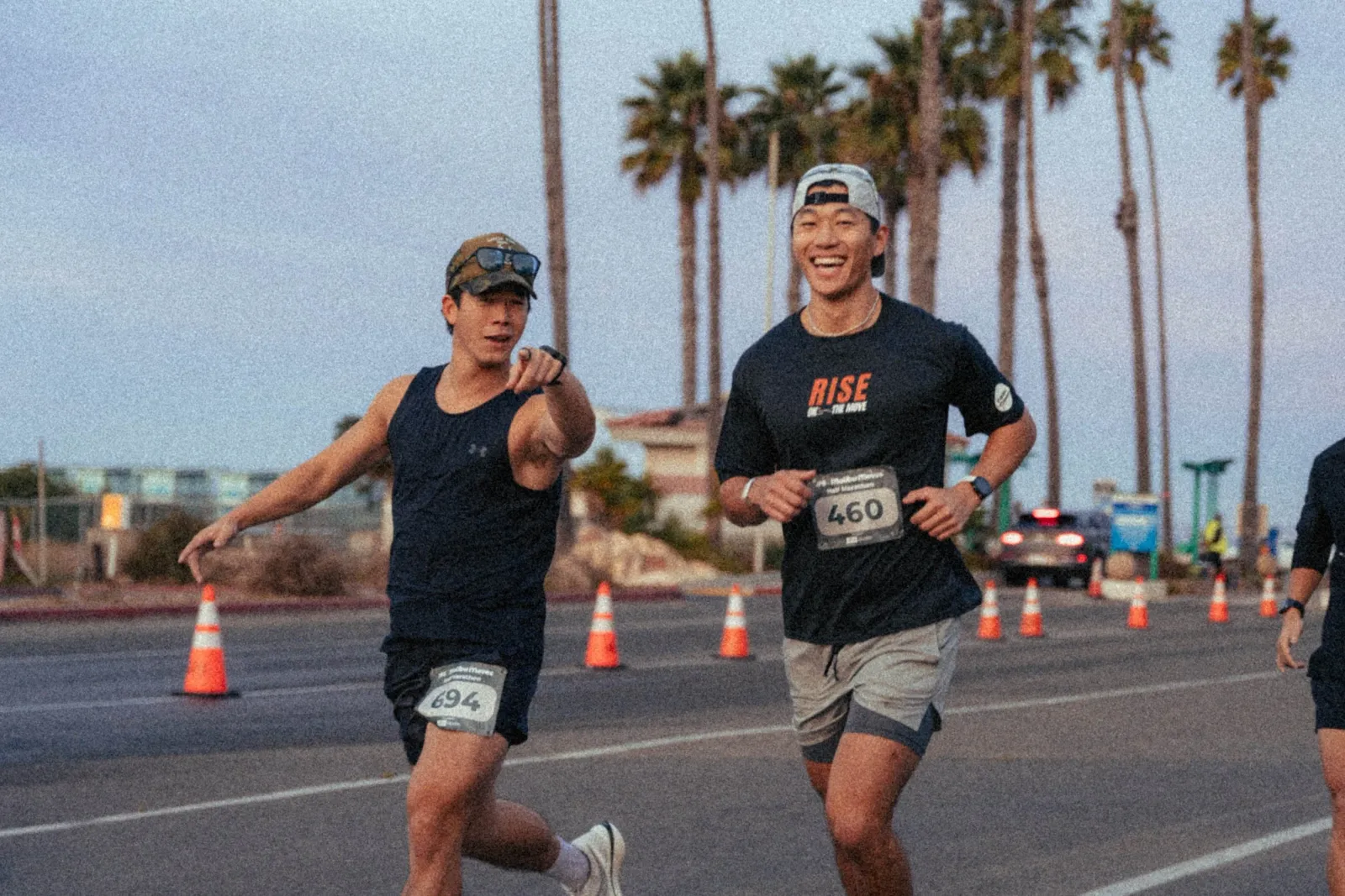 Two male runners smiling and running on a road lined with palm trees, one pointing towards the camera.