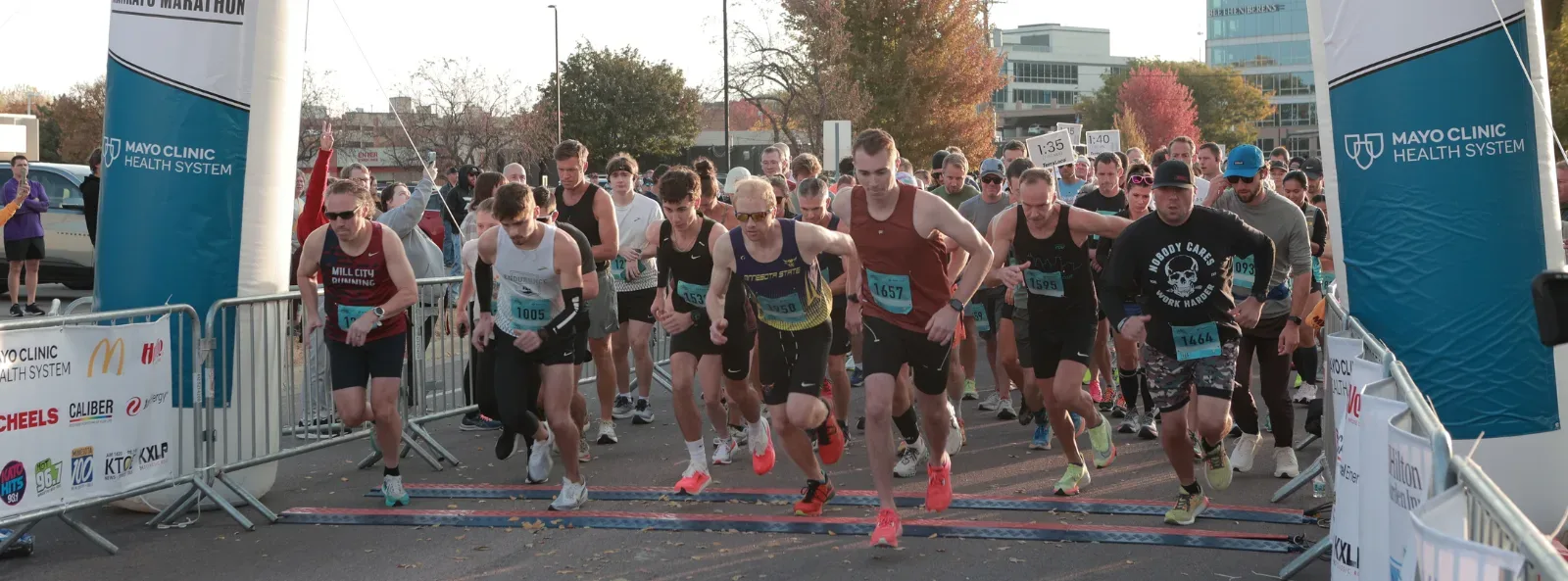 Runners jostling at the start line of a half marathon, with banners and barriers in the foreground and spectators in the background.