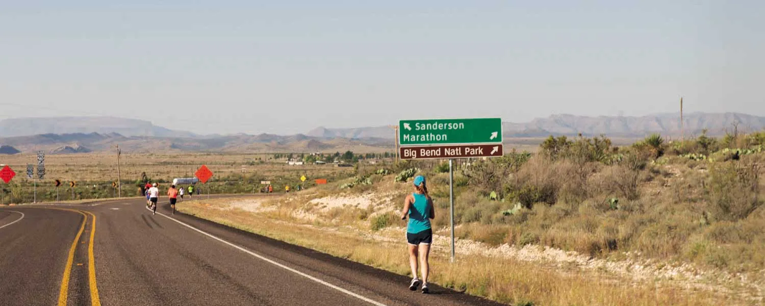 A female runner in a teal tank top and black shorts runs on a road with a green sign indicating 'Sanderson Marathon' and 'Big Bend Natl Park' in the background. Other runners are visible further down the road.