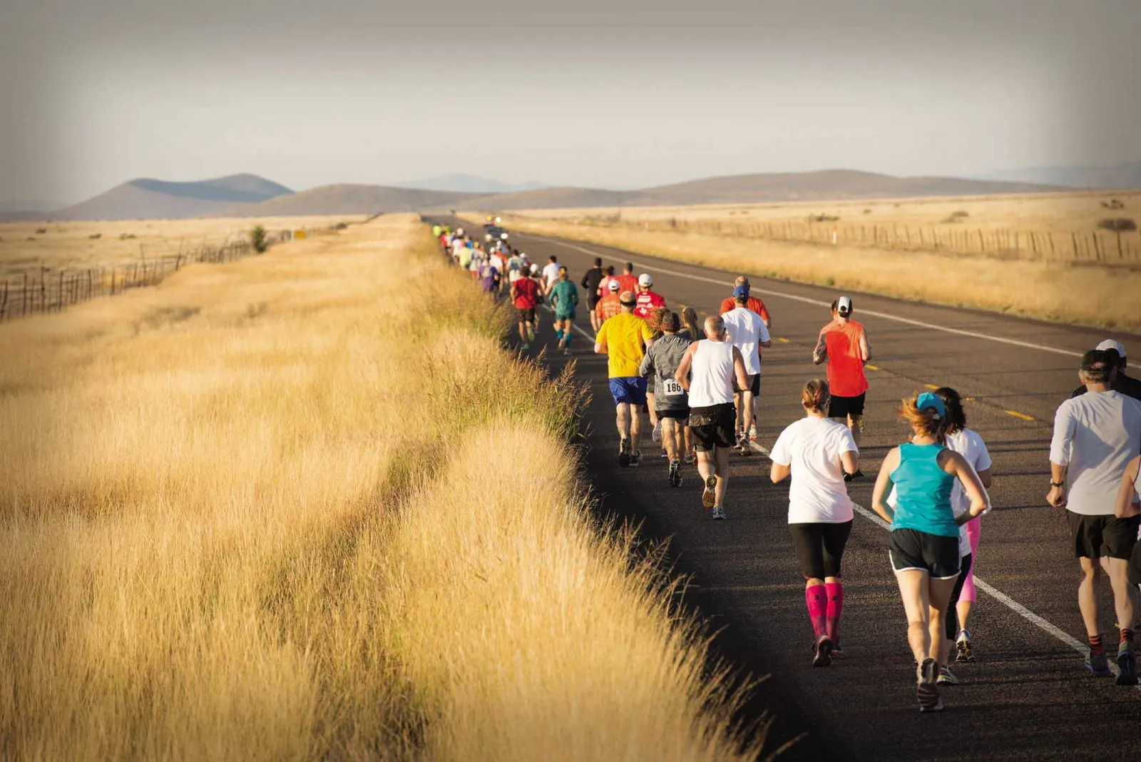 Runners on a road with dry grass fields and distant hills under a hazy sky.