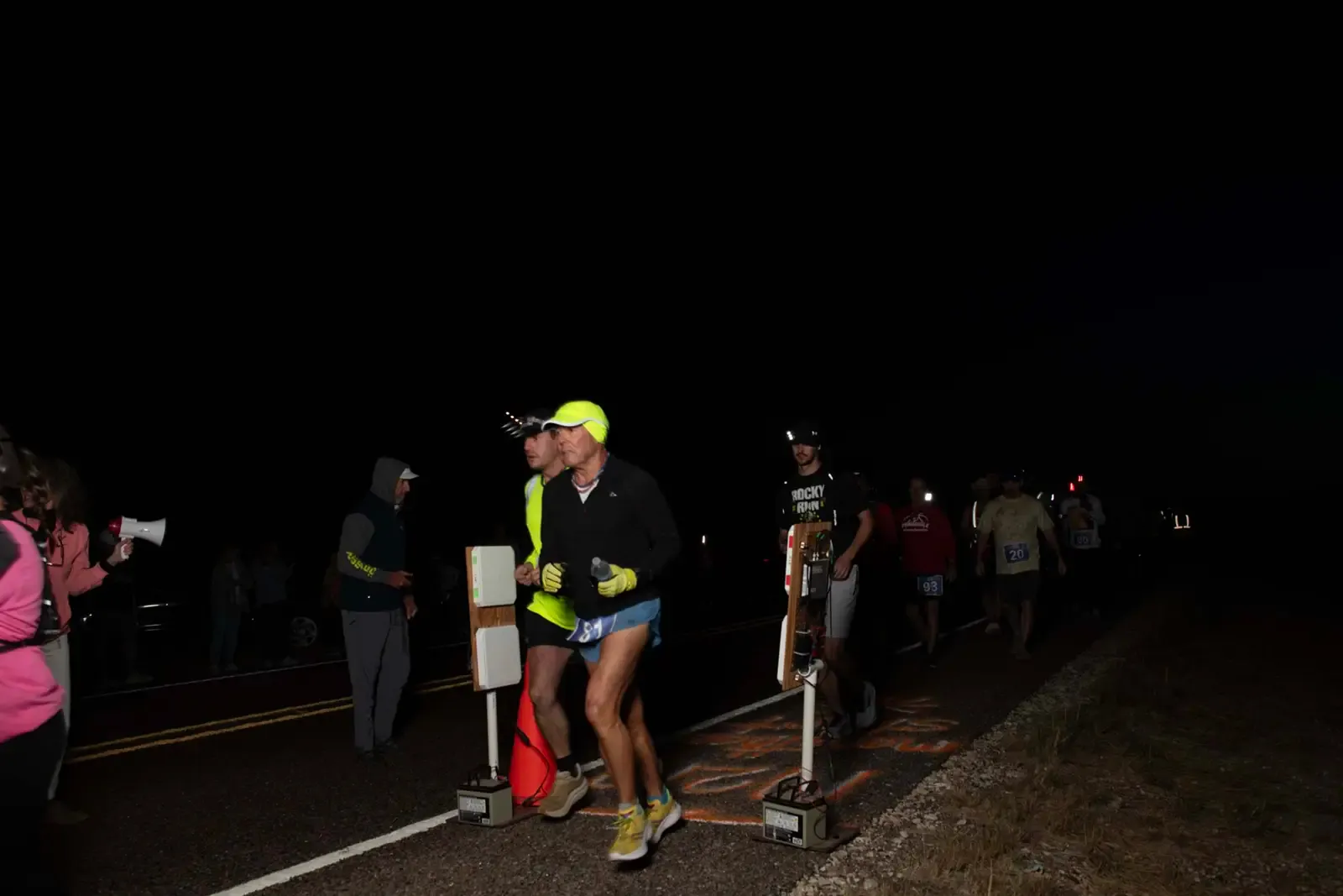 Runners starting a race at night, with some spectators and race officials visible.