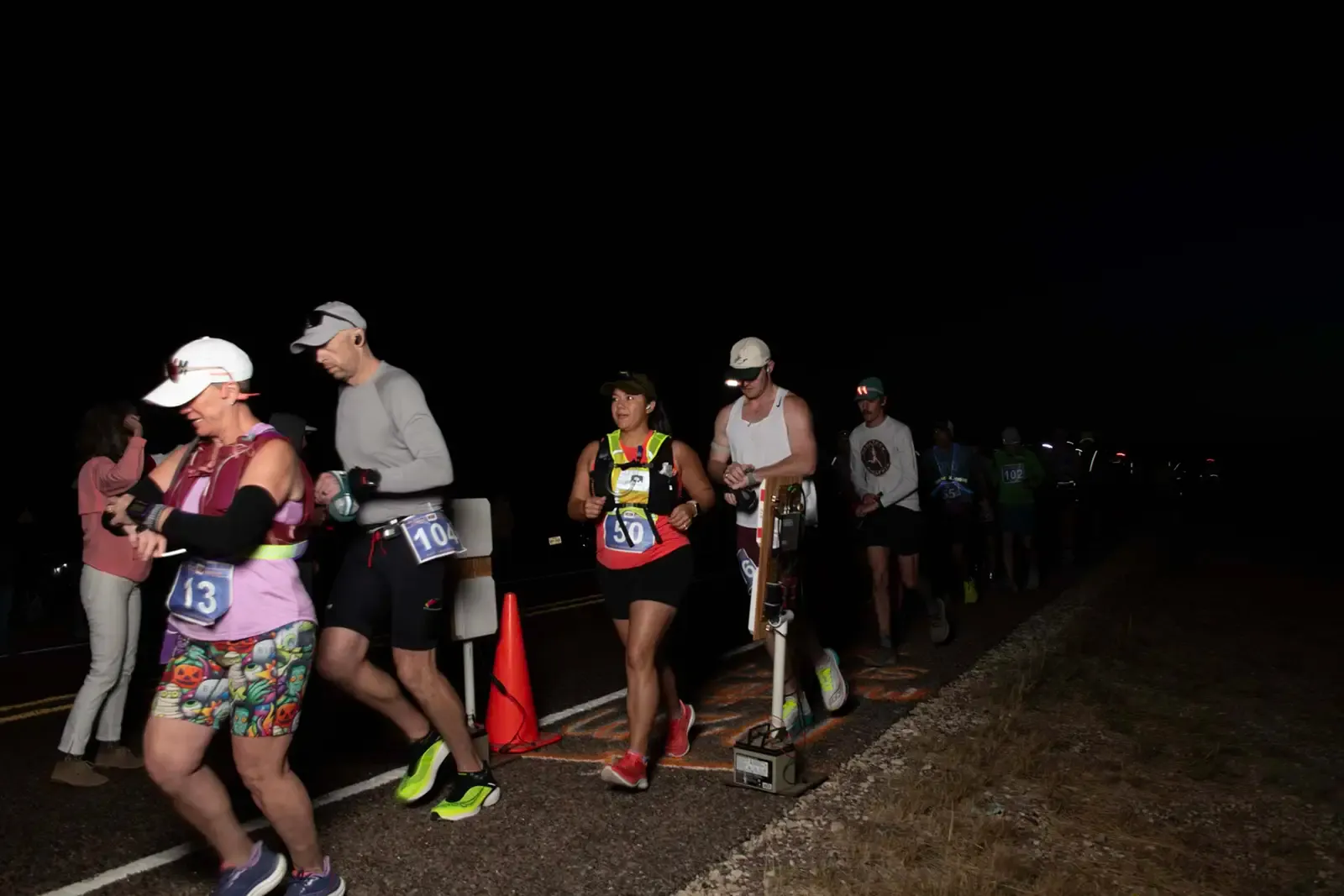 Runners in a race at night, some wearing headlamps, with bib numbers visible.
